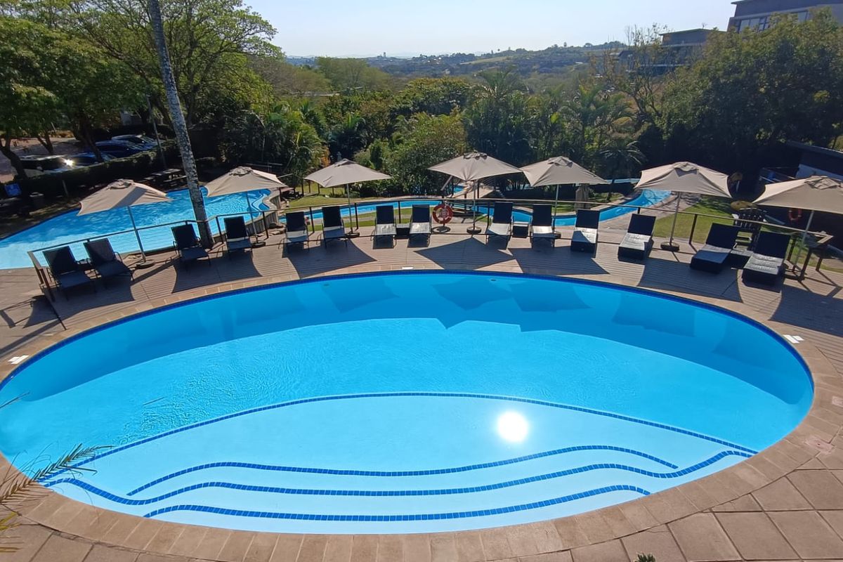 A well-maintained outdoor swimming pool area featuring a large oval pool with integrated steps, surrounded by a paved deck and scenic landscape views.
