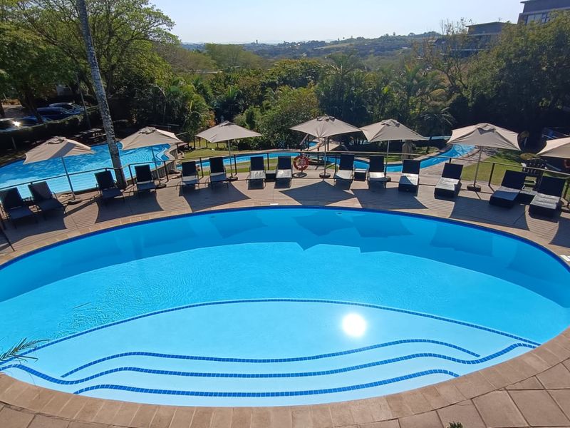 A well-maintained outdoor swimming pool area featuring a large oval pool with integrated steps, surrounded by a paved deck and scenic landscape views.