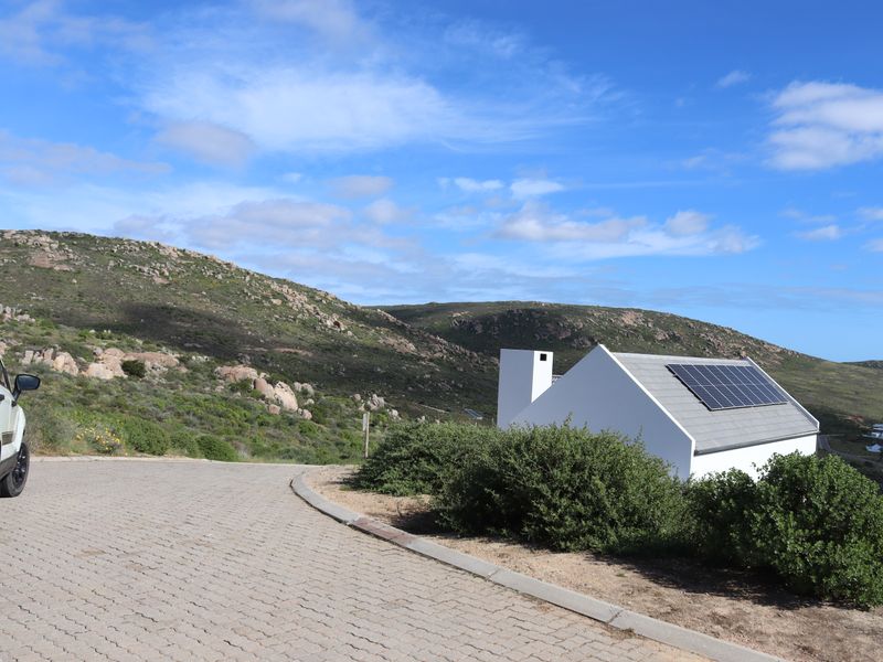 A view of a residential property featuring a white facade, solar panels on the roof, and a paved driveway set against a hilly landscape.