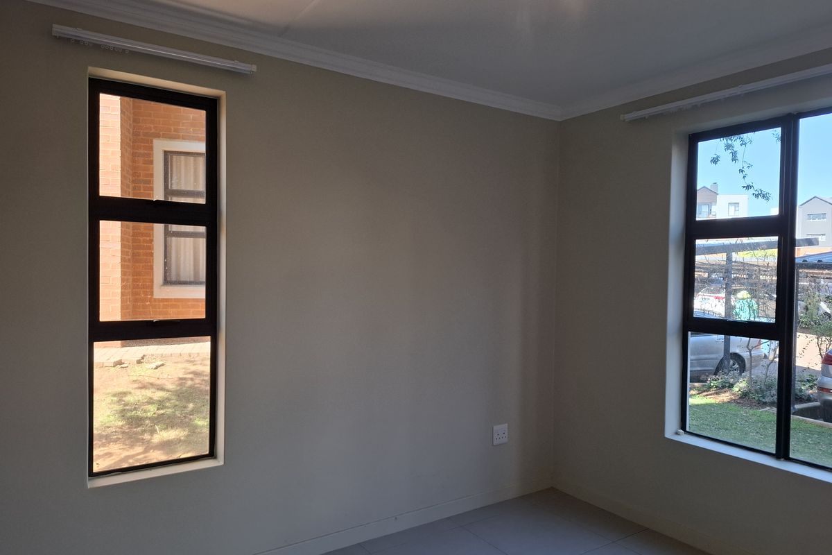 A bright, empty bedroom featuring neutral wall tones, tiled flooring, and large aluminium-framed windows providing natural light and views of the surrounding complex.