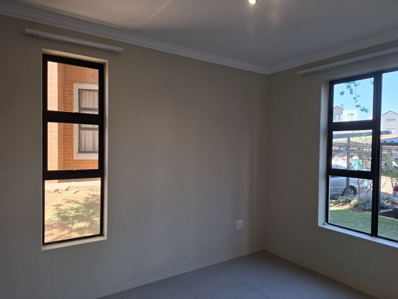 A bright, empty bedroom featuring neutral wall tones, tiled flooring, and large aluminium-framed windows providing natural light and views of the surrounding complex.