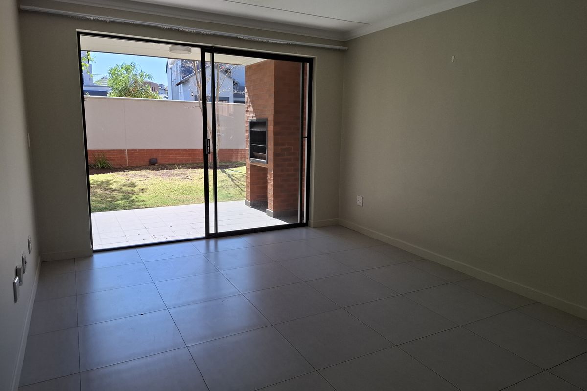 An empty living room featuring large sliding glass doors that open onto a paved patio with a built-in brick braai and garden view.