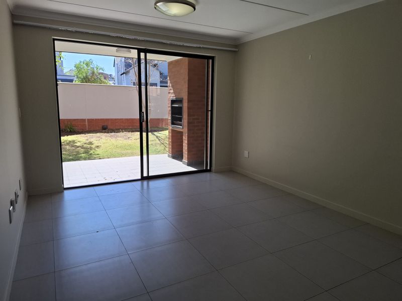 An empty living room featuring large sliding glass doors that open onto a paved patio with a built-in brick braai and garden view.