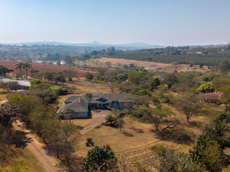Aerial view of a residential property featuring a tiled roof home, paved driveway, and expansive surrounding grounds in a rural landscape setting.