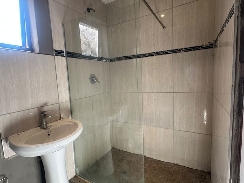 A compact bathroom featuring a pedestal basin, a walk-in shower with a glass screen, and neutral wall tiling.