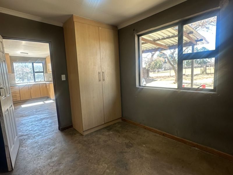 An empty bedroom featuring concrete flooring, a built-in cupboard, and an aluminium window, with a view through to the adjacent kitchen area.