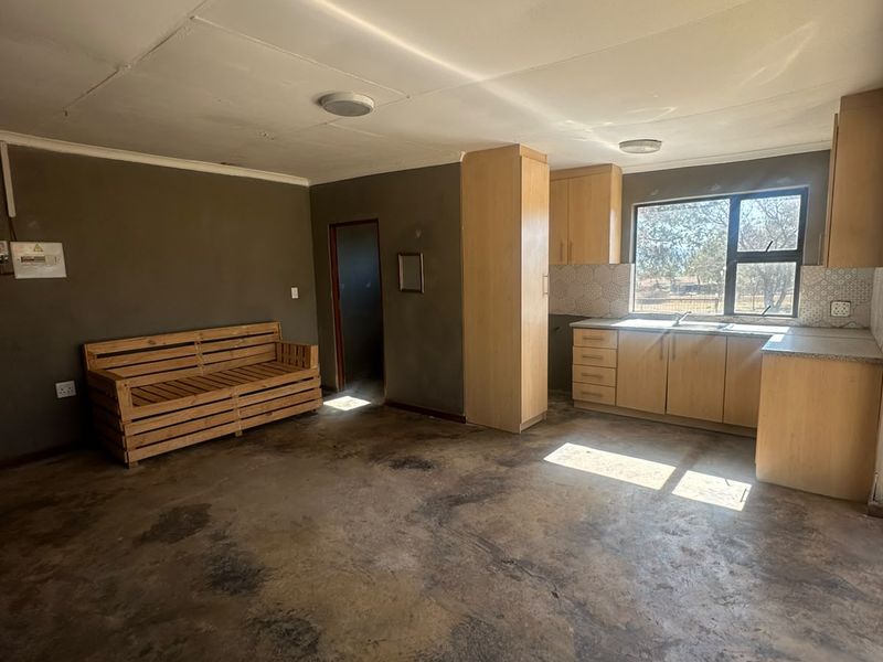 Open plan living area featuring a compact kitchen with light wood cabinetry, laminate countertops, and a tiled backsplash, with worn concrete flooring throughout.
