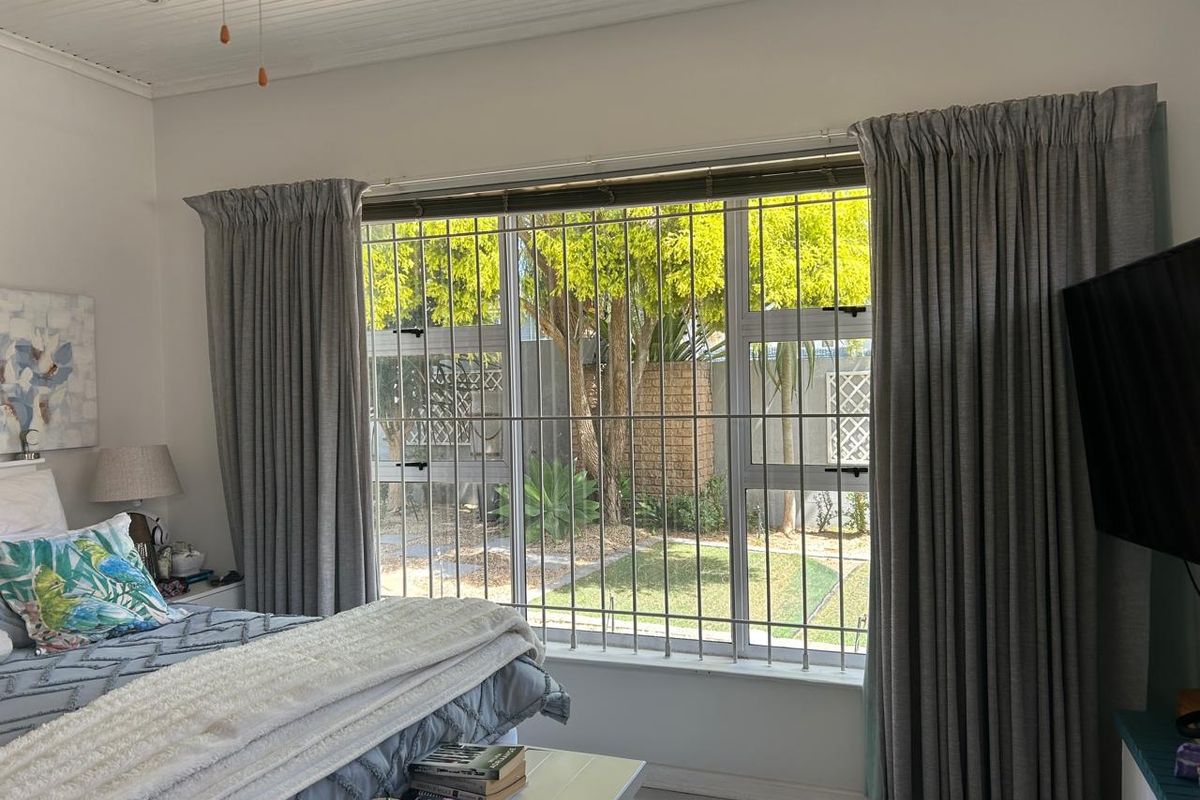 A bright bedroom featuring a large window with security bars, overlooking a private garden area, complemented by a classic tongue and groove ceiling.