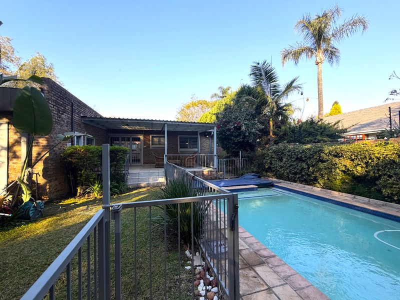 Rear exterior view featuring a swimming pool with safety fencing, a covered patio area, and established garden landscaping.
