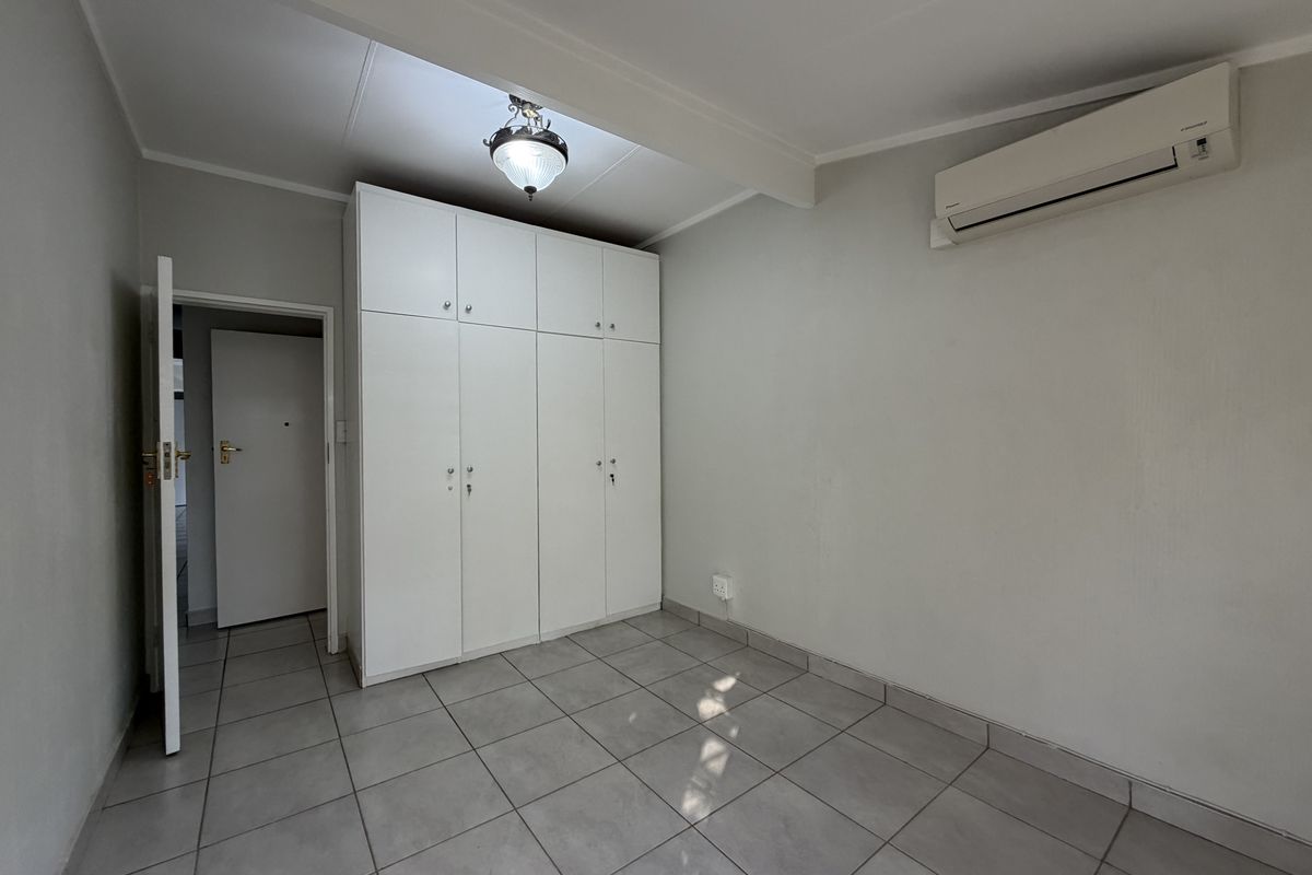 A clean, empty bedroom featuring neutral tiled flooring, built-in white cupboards, and a wall-mounted air conditioning unit.