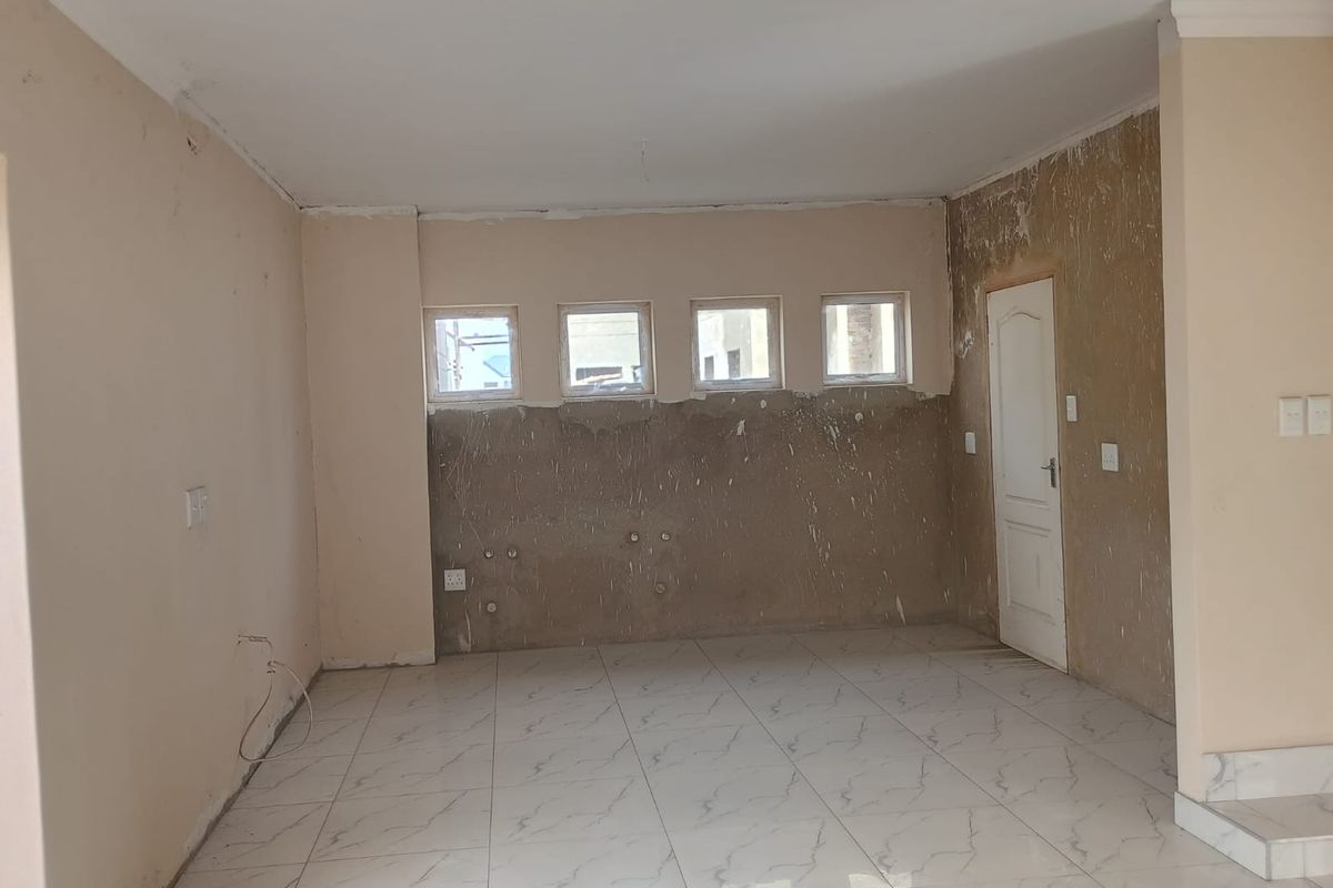 An empty, unfinished kitchen area featuring marble-look tiled flooring, plumbing connections, and four windows, currently requiring wall finishing and final installation of cabinetry.