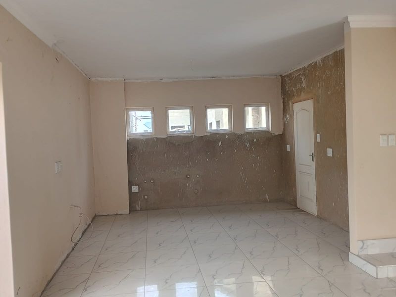 An empty, unfinished kitchen area featuring marble-look tiled flooring, plumbing connections, and four windows, currently requiring wall finishing and final installation of cabinetry.