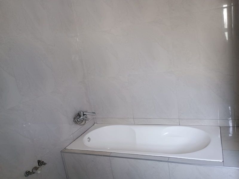 A clean, modern bathroom featuring a built-in bathtub with wall-mounted chrome fixtures and neutral large-format wall tiles.