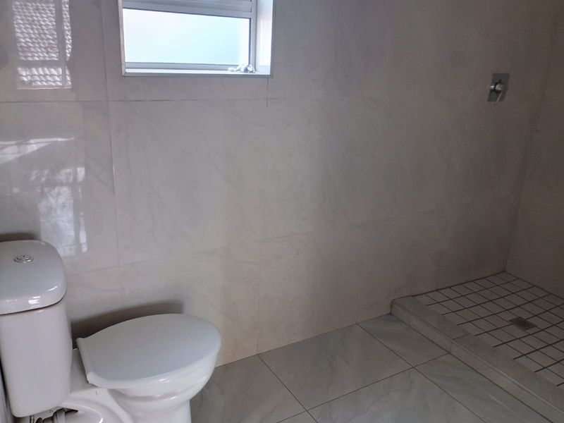 A clean, modern bathroom featuring neutral wall and floor tiles, a white toilet, and a walk-in shower area with a tiled base.