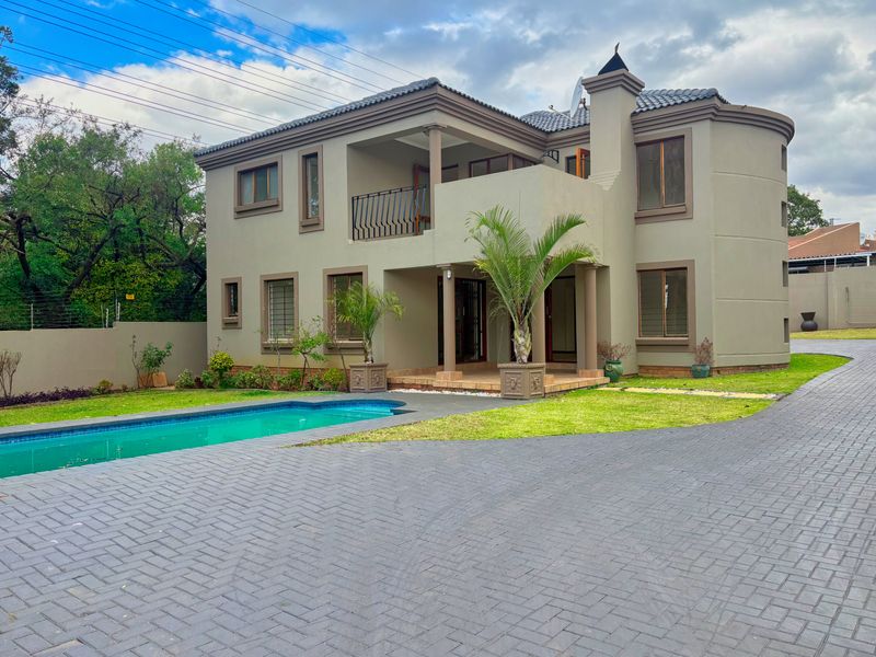 A two-story residential property featuring a swimming pool, paved driveway, and a balcony, set within a landscaped garden with perimeter security.