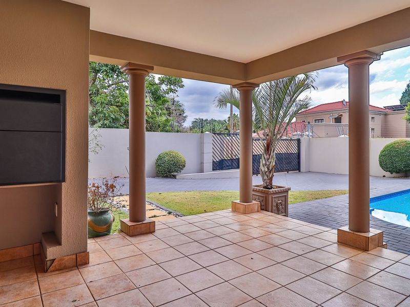 Covered patio area featuring a built-in braai, tiled flooring, and support columns, overlooking a private swimming pool and paved driveway.