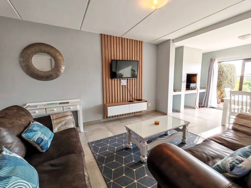Open-plan living area featuring a modern timber-slat television wall, floating media console, and a built-in indoor braai, leading out to a patio.