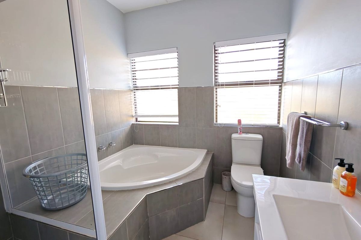 A well-maintained bathroom featuring a corner bathtub, tiled walls and flooring, a vanity basin, and dual windows with venetian blinds.