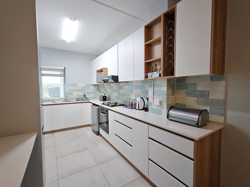 Modern galley-style kitchen featuring white cabinetry, wood-tone accents, stone countertops, a tiled backsplash, and integrated cooking appliances.