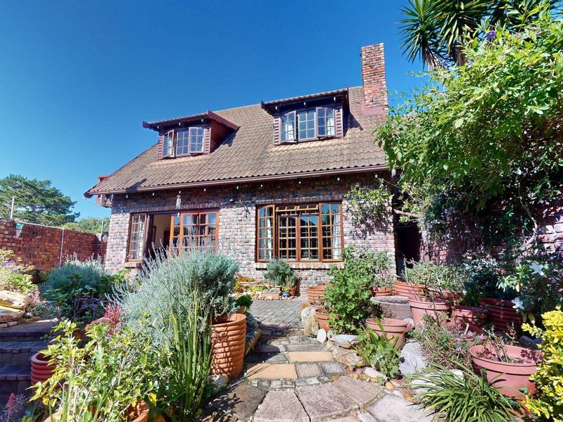 A charming rear exterior view of a brick townhouse featuring a tiled roof, dormer windows, a brick chimney, and a stone-paved patio area.