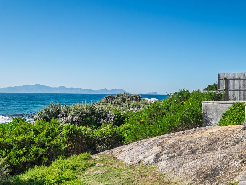 A scenic coastal view featuring natural rock formations, lush coastal vegetation, and a clear ocean horizon with distant mountain ranges.