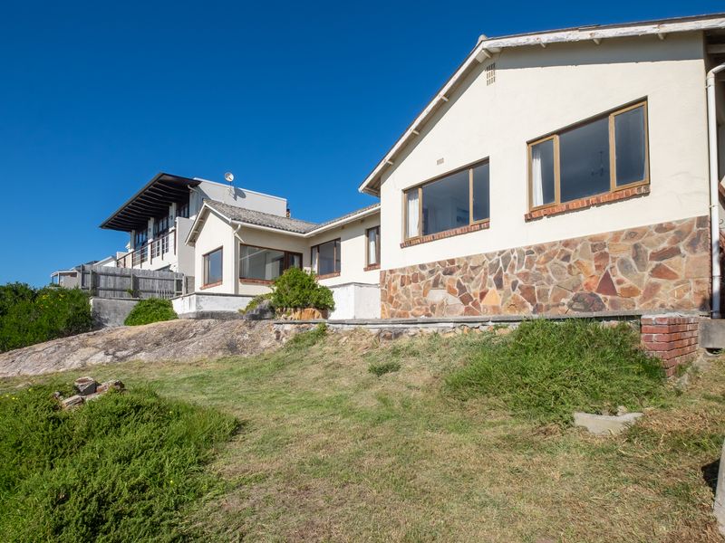 Rear view of the property featuring stone cladding, timber window frames, and a sloped garden area with natural rock formations.