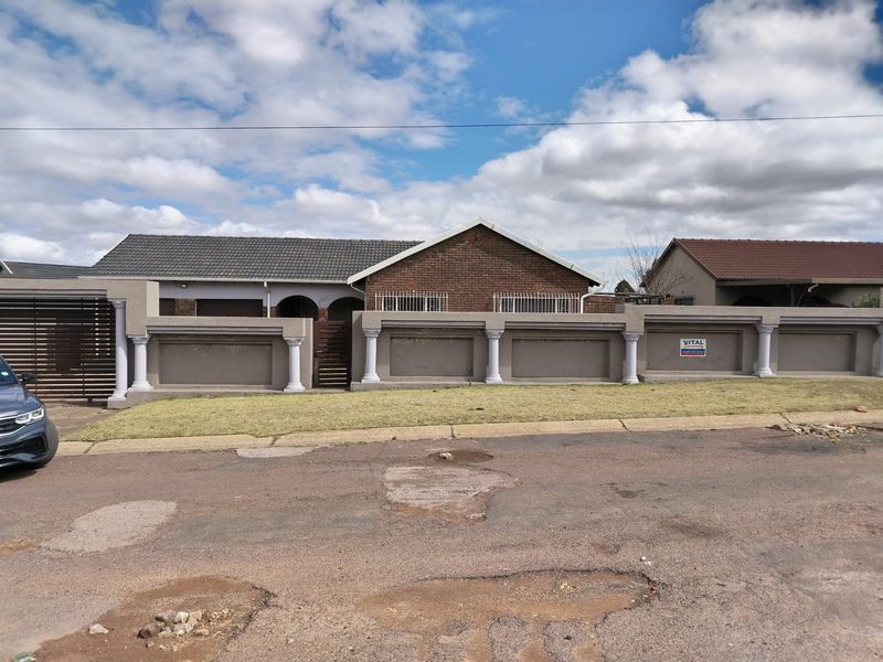 Front view of a single-story brick house featuring a prominent perimeter wall with decorative pillars and a secure gated entrance.