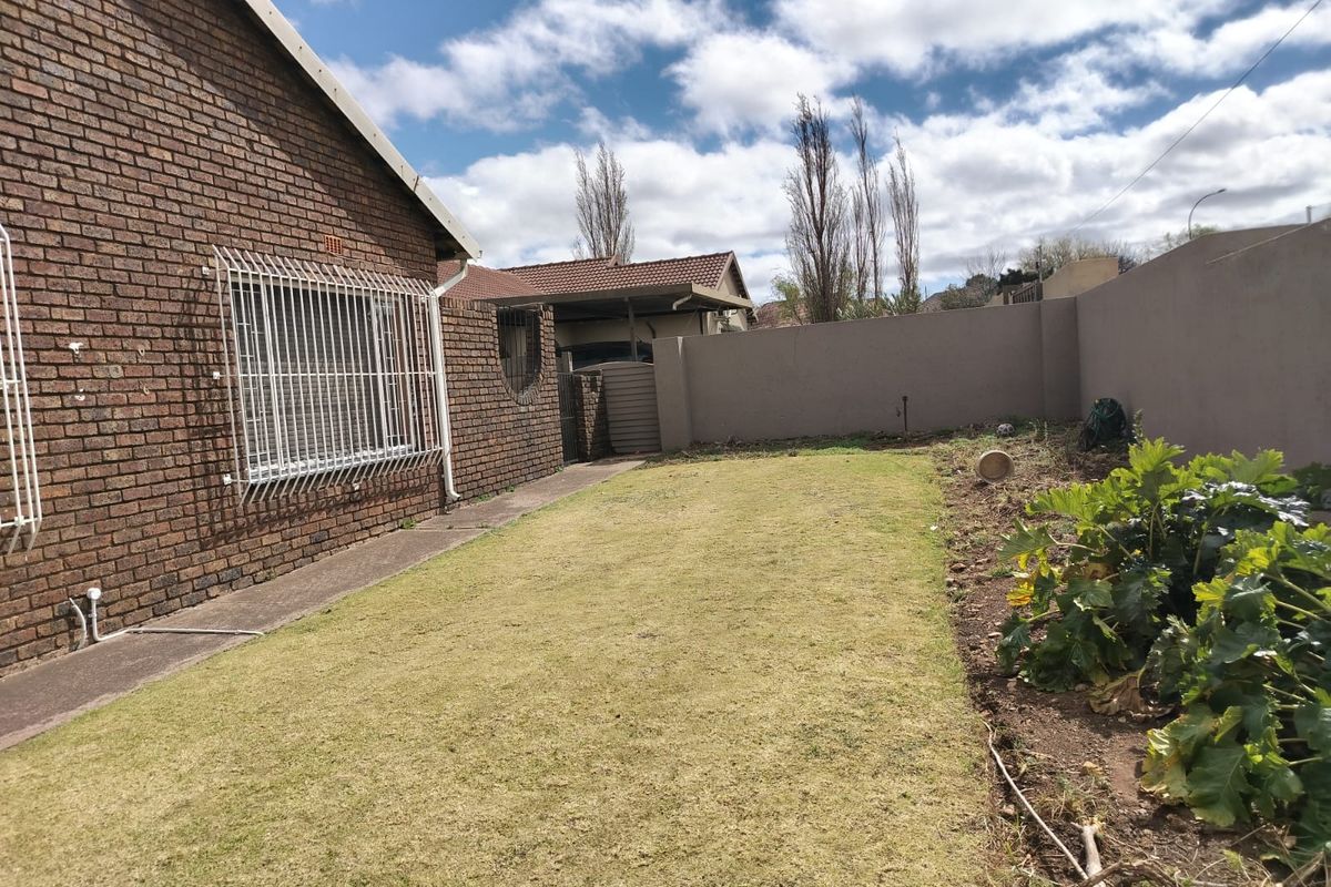 A rear exterior view of the property featuring a brick facade with security bars, a lawn area, and a boundary wall.