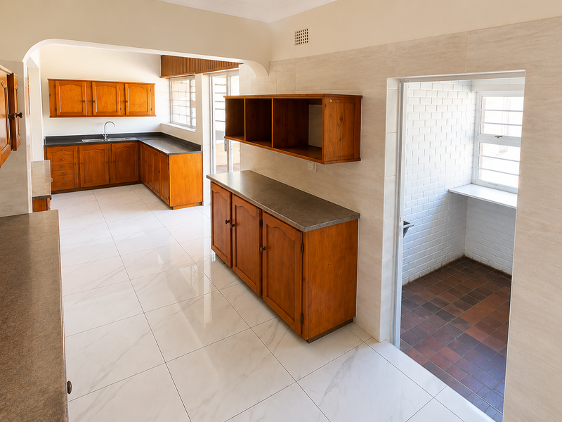 A spacious, empty kitchen featuring wooden cabinetry, laminate countertops, and light-colored floor tiles, with an adjacent utility area and arched transition.
