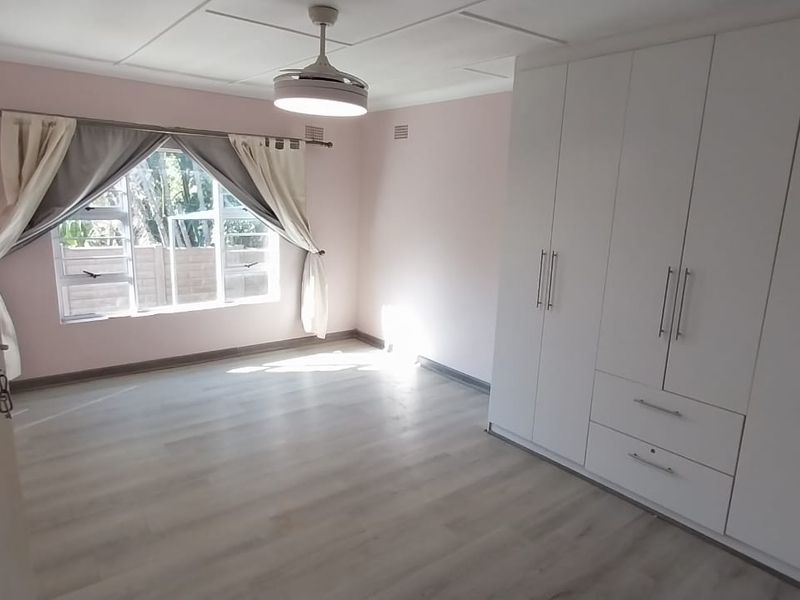 A bright, empty bedroom featuring modern laminate flooring, built-in white cupboards, and a ceiling fan, offering a clean and neutral space.