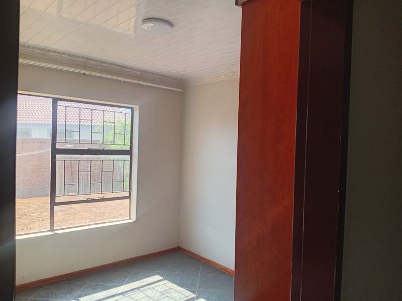 A vacant bedroom featuring tiled flooring, a window with security burglar bars, and decorative ceiling cornicing, viewed through a wooden door frame.
