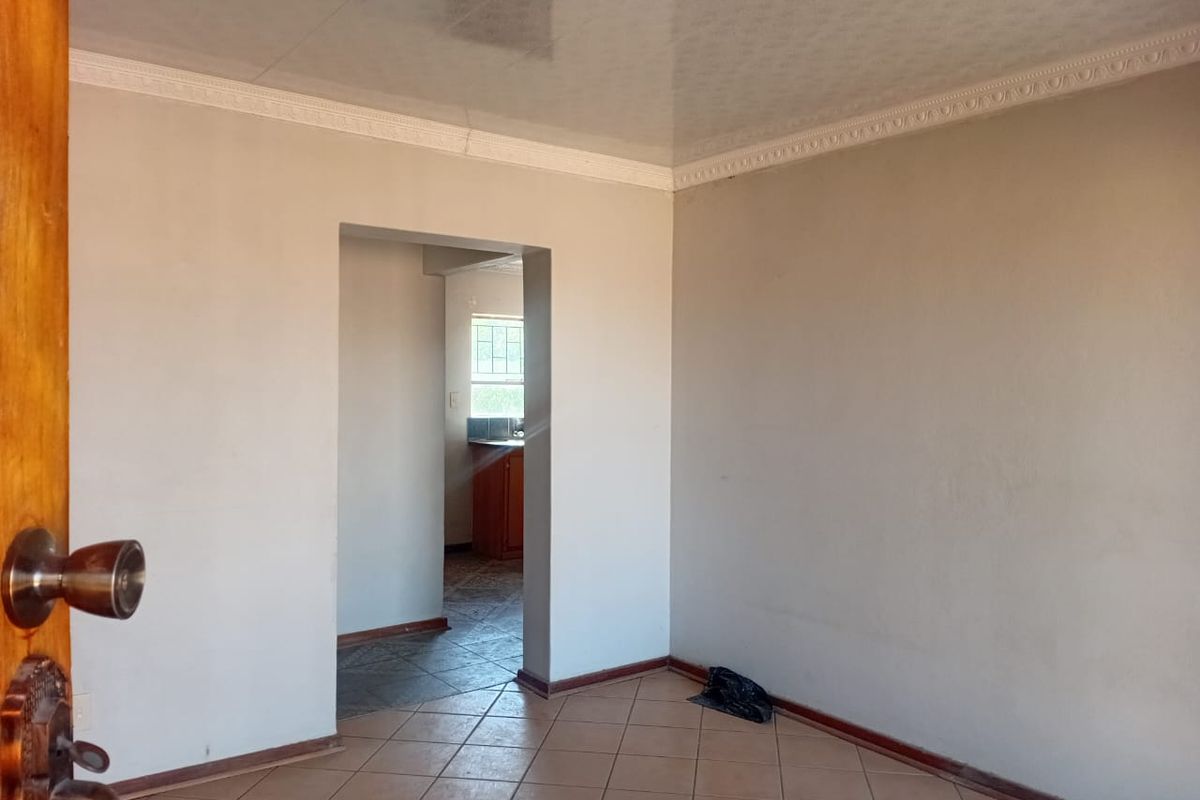 An empty living room featuring tiled flooring and decorative ceiling cornicing, with a doorway leading to an adjacent kitchen area.