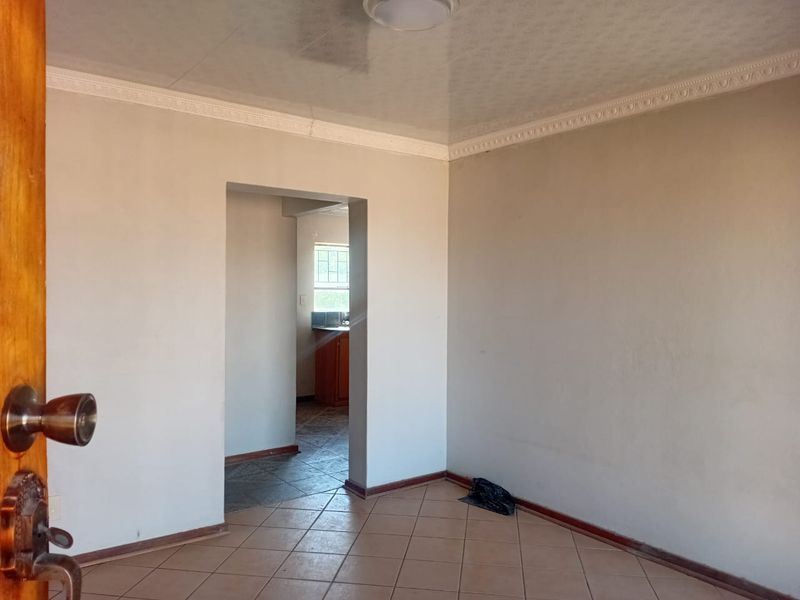 An empty living room featuring tiled flooring and decorative ceiling cornicing, with a doorway leading to an adjacent kitchen area.