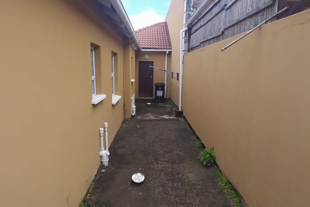 A narrow rear exterior service area featuring concrete paving, boundary walls, and external plumbing fixtures, providing access to the rear entrance.