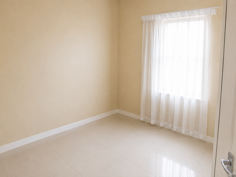 A bright, empty bedroom featuring neutral-toned walls, large format floor tiles, and a window fitted with a curtain rail.