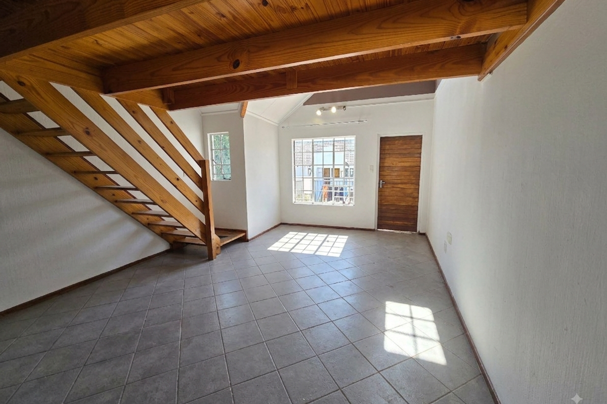 An empty living room featuring tiled flooring, exposed wooden ceiling beams, and a wooden staircase leading to an upper level.