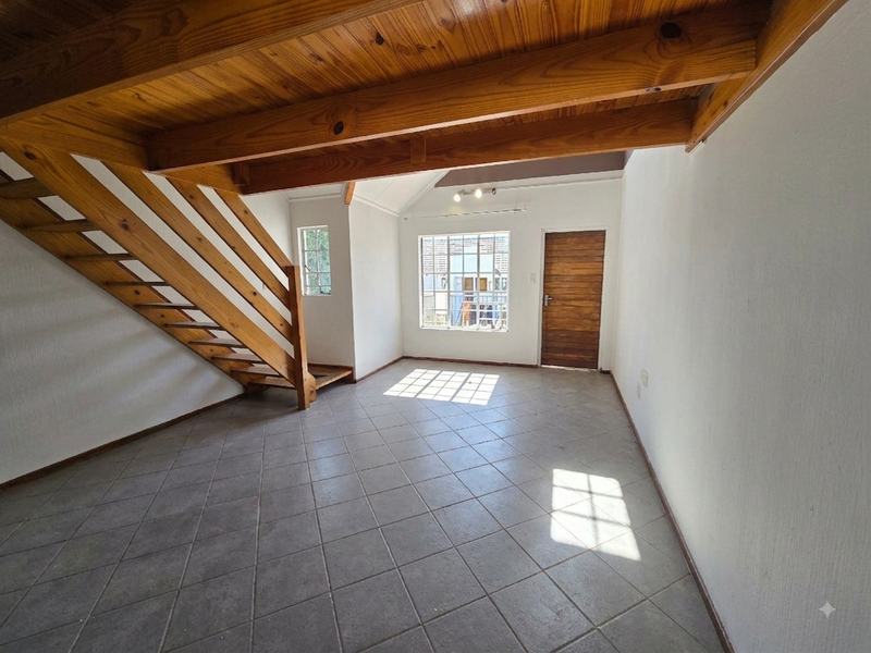 An empty living room featuring tiled flooring, exposed wooden ceiling beams, and a wooden staircase leading to an upper level.