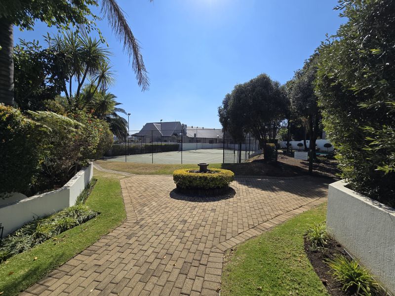 A well-maintained communal tennis court area featuring a paved walkway, landscaped garden beds, and perimeter fencing within the townhouse complex.