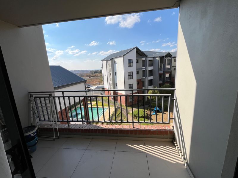 A covered balcony featuring tiled flooring and a metal balustrade, offering views over the apartment complex grounds and swimming pool area.
