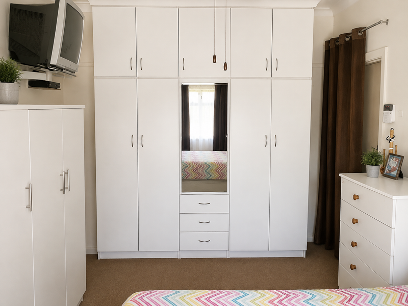 A bedroom featuring a large white built-in wardrobe with integrated mirror and drawer storage, providing ample organized clothing space.