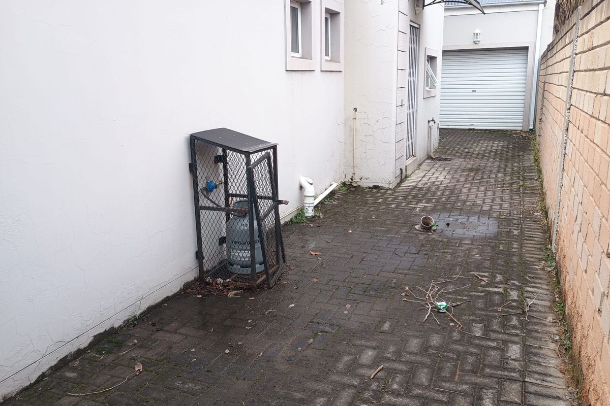 Rear exterior area featuring paved flooring, a gas cylinder storage cage, and access to the garage, showing signs of wear and maintenance requirements.