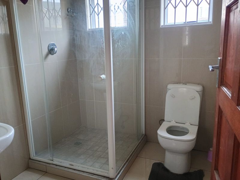 A functional bathroom featuring a glass-enclosed shower, toilet, and neutral wall and floor tiling with security bars on the windows.