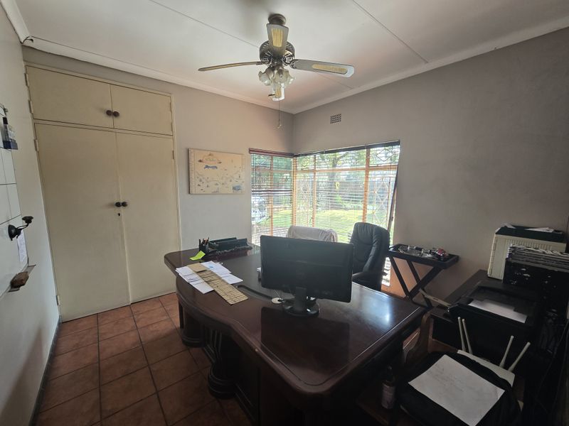 A functional home office featuring built-in storage cupboards, tiled flooring, and a ceiling fan, with large windows providing natural light.