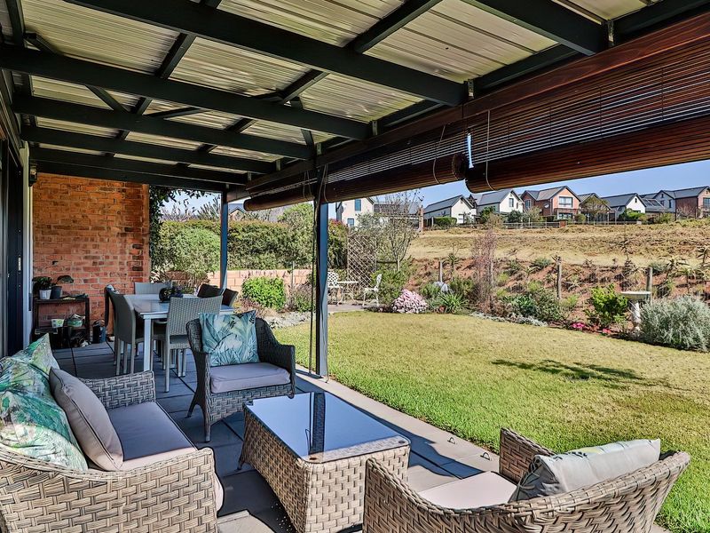 A covered outdoor patio featuring paved flooring, exposed brickwork, and a metal roof structure, overlooking a landscaped garden and residential neighborhood.
