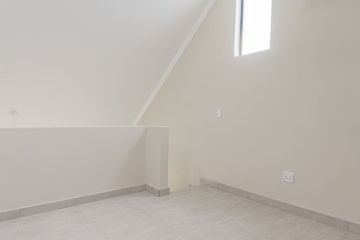 An empty, neutral-toned bedroom featuring tiled flooring, a sloped ceiling with crown molding, and a small window providing natural light.
