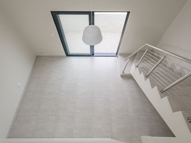 A bright, modern open-plan living area featuring neutral tiled flooring, a sleek stainless steel staircase railing, and large sliding glass doors leading outdoors.