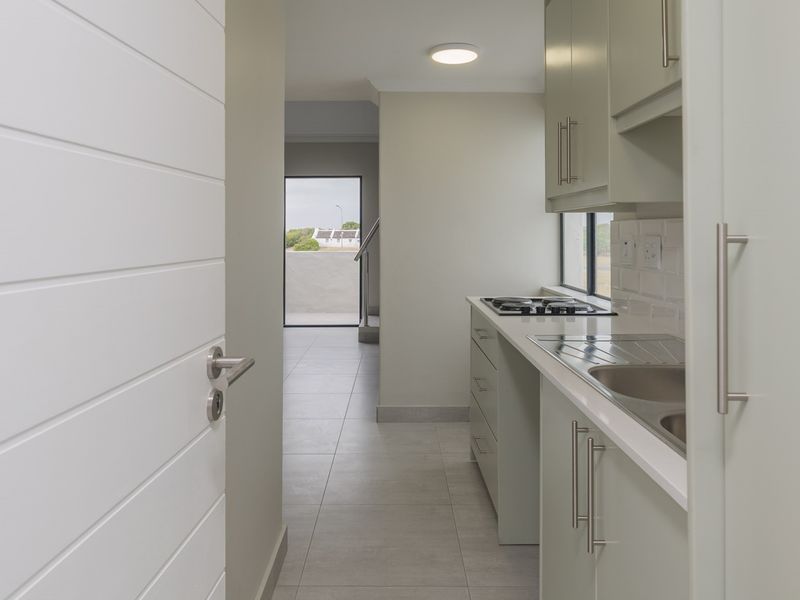 A modern, clean kitchen featuring light-toned cabinetry, a gas hob, stainless steel sink, and neutral tiled flooring, leading to an open living area.
