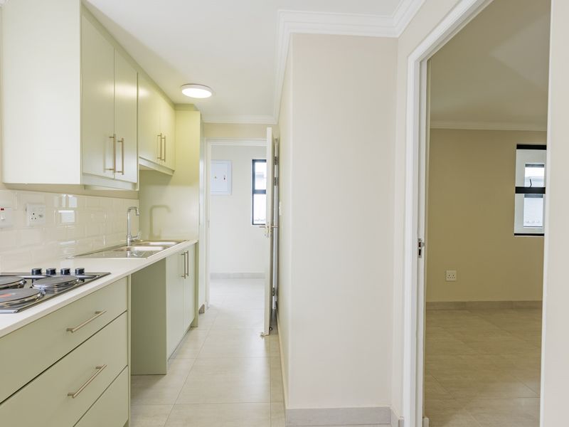 A clean, modern kitchen featuring light green cabinetry, a gas hob, subway tile backsplash, and tiled flooring, leading into an adjacent living area.