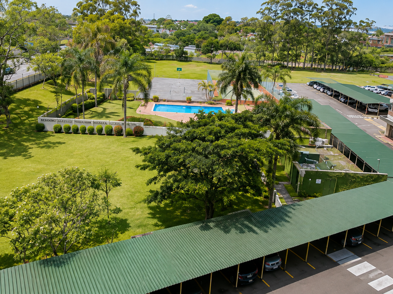 Aerial view of the apartment complex grounds featuring a swimming pool, tennis court, landscaped gardens, and extensive covered parking facilities.