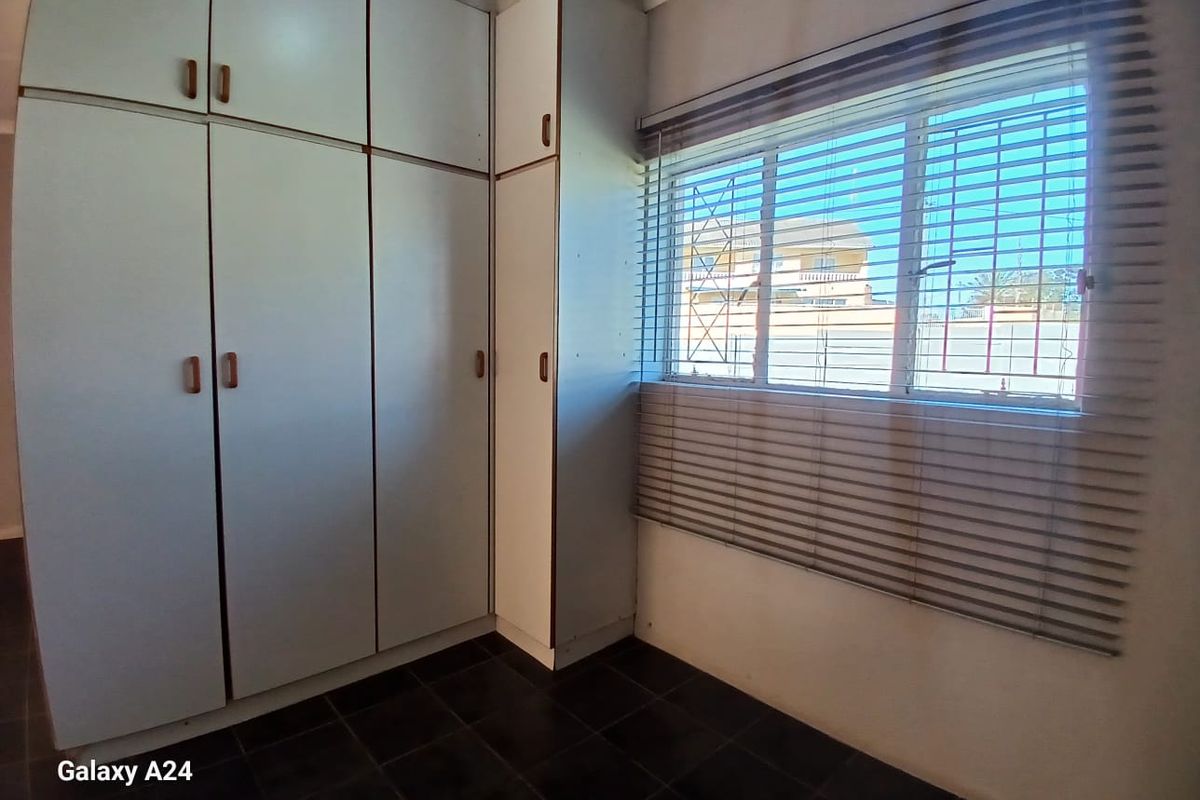A clean, empty bedroom featuring built-in cupboards, dark tiled flooring, and a window fitted with security bars and venetian blinds.